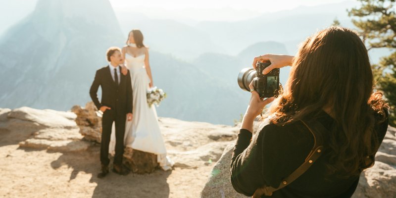A wedding photographer photographing a bride and groom during an intimate outdoor elopement on a rocky overlook with mountains in the background.