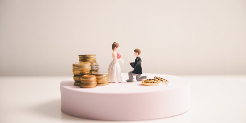 Bride and groom cake topper beside stacked coins and wedding rings symbolizing wedding expenses and who pays for a wedding.
